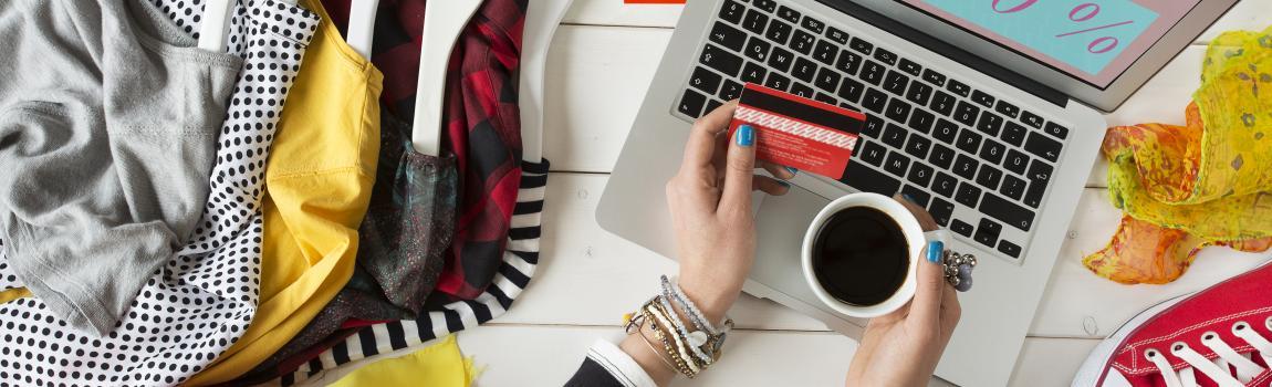 iStock.com/ seb ra chaotischer Schreibtisch mit Lap Top auf Online-Shopping-Seite, daneben viele Kleider, Kaffee, Konsumgüter